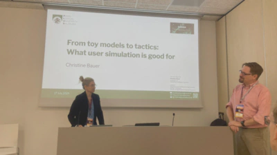 Photo of Christine Bauer (left-hand side) in front of a slide projection saying ‘From toy models to tactics: What user simulation is good for’; next to Philipp Schaer introducing her (on the right-hand side)